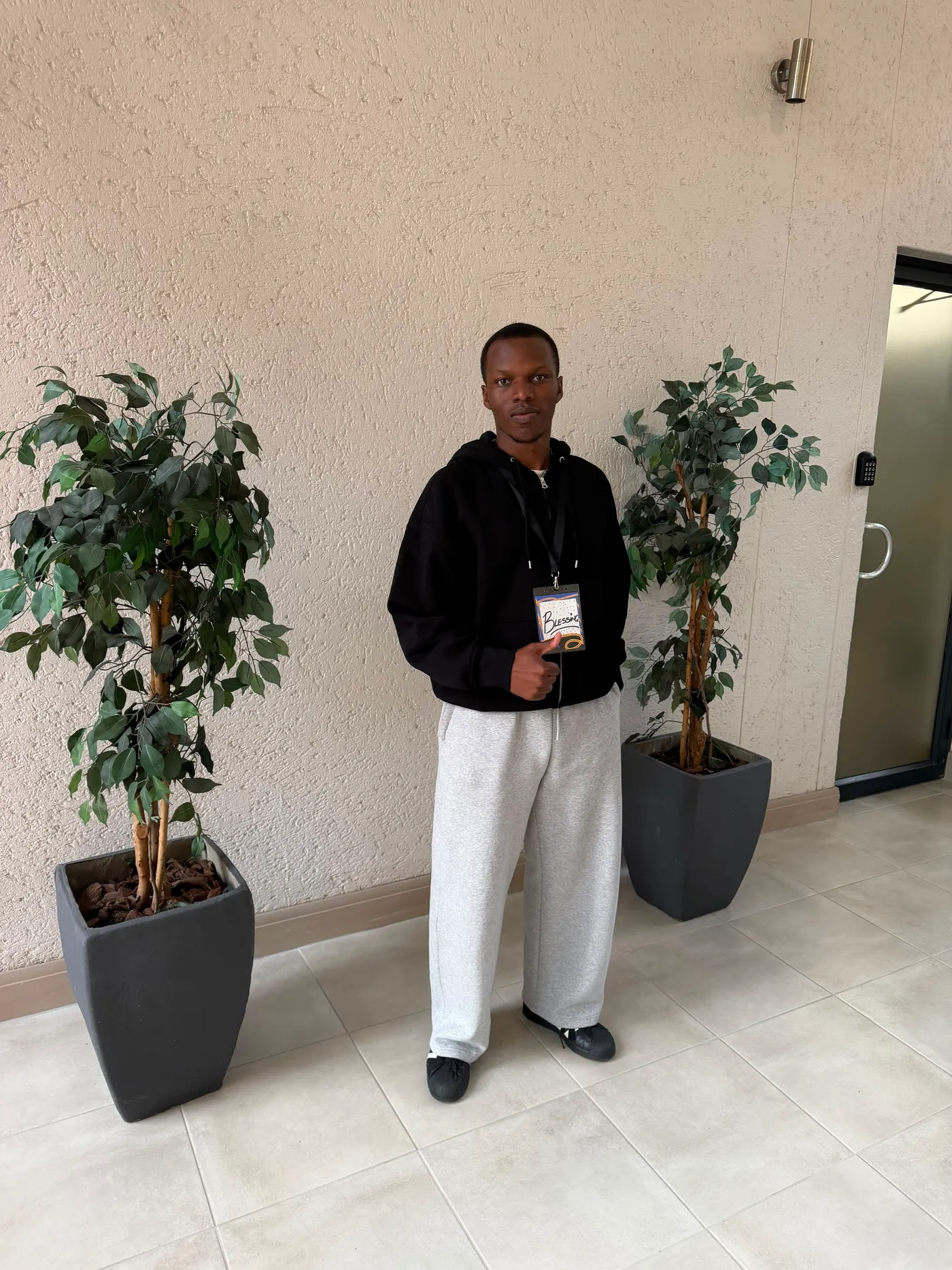 Blessing standing between two potted ficus trees with a name badge in his hand
