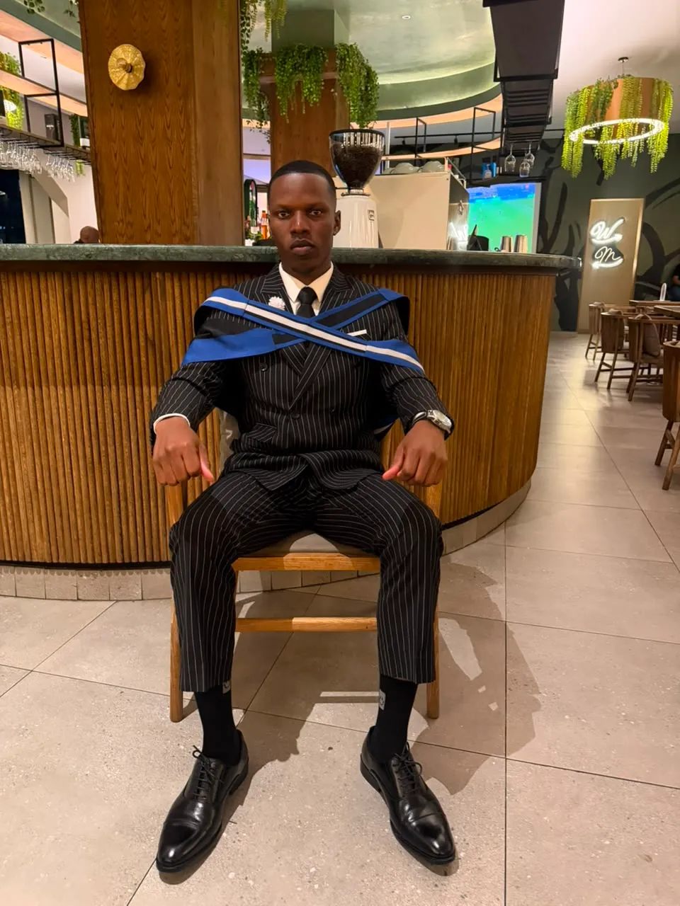 Blessing seated in a pinstriped suit with a graduation sash across his chest, hands resting on his knees