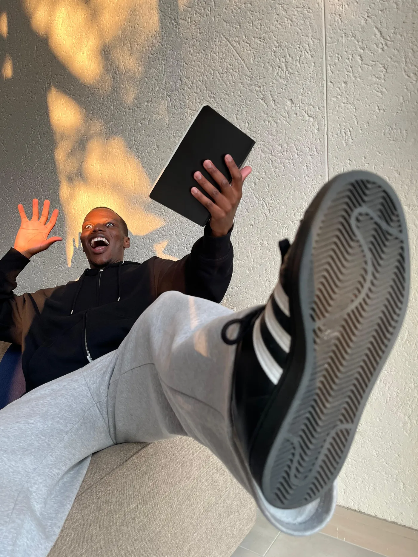 Blessing sprawled back on a couch mid-laugh, a notebook held up in one hand and the other thrown open, one sneaker kicked toward the camera and leaf shadows falling across the wall behind him
